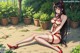 A woman in red lingerie and fishnet stockings sits on a stone path surrounded by potted plants.